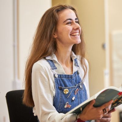 A girl with brown hair and wearing blue dungarees and a white shirt holds out a book. She has a big smile across her face.